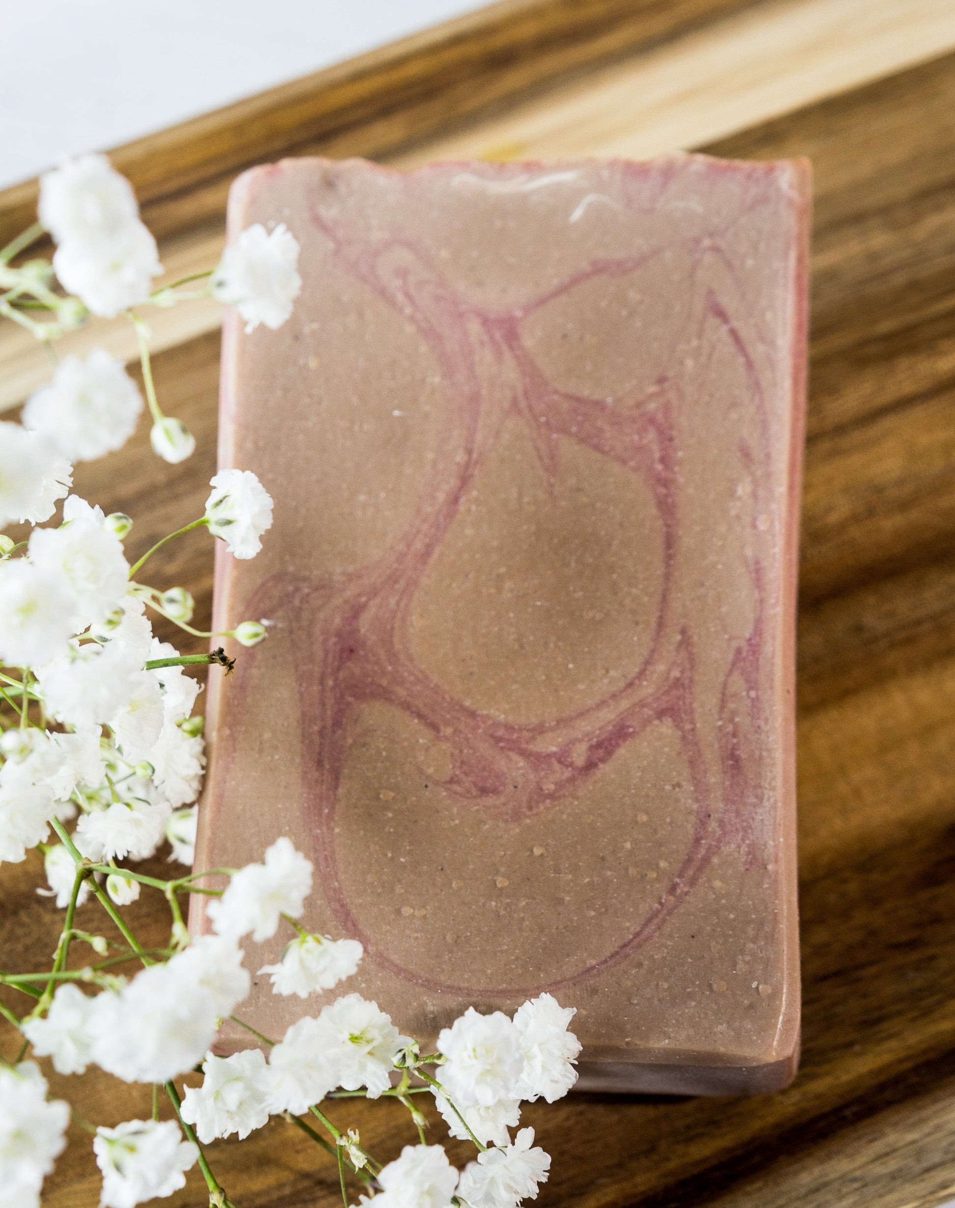 Amber and Oud with Pumice bar soap with pink and tan swirls set on a wood stand with baby's breath flowers..