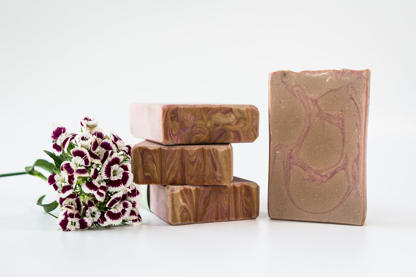 Amber and Oud with Pumice bar soap with pink and tan swirls, one standing and three stacked. Dark pink and white flowers next to the stack of three. 